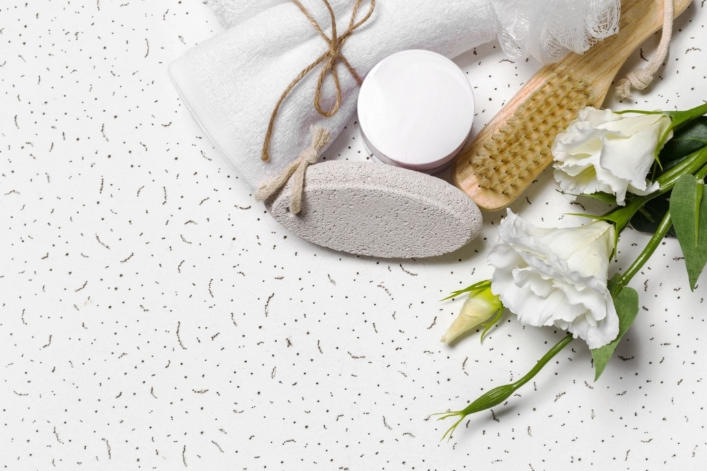 Pummace stone, bath supplies, and flowers sitting on white quartz countertop with brown speckles.